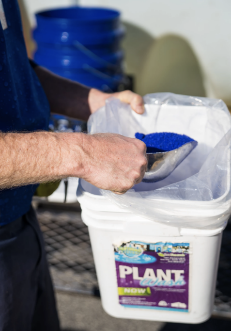 A person is scooping blue granular substance from a white plastic container labeled "PLANT" with other small text on the label. The person is wearing a blue shirt, and there are buckets and industrial equipment in the background.